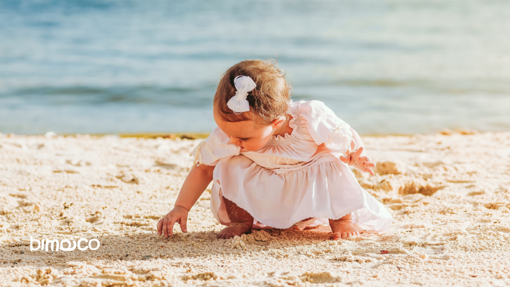 Baby in a pale pink dress kneels in the sand at the beach, digging with tiny fingers as the sea glows in the background.