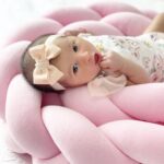 Close-up of a newborn baby girl resting on a soft pink pillow, wearing a floral outfit and peach bow headband.