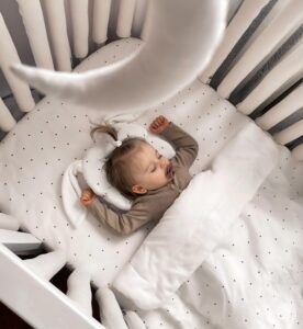 Infant sleeping on white bedding with small black dots in a crib, pacifier in mouth.