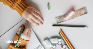 Child's hands write in a grid notebook on a white desk, surrounded by colored pencils and an eraser case with a pink lid (bimico logo)