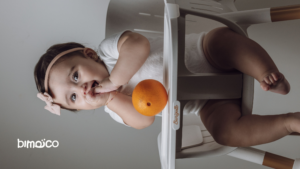 Baby sitting in a high chair, wearing a pink headband and white outfit, reaching for an orange on the tray.