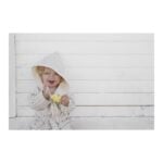 Baby in a striped hooded robe sits against a white wooden wall, crying while holding a piece of pineapple.