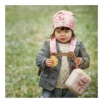 Young child in a pink hat and plaid jacket outdoors, holding a cookie and a pastel lunchbox tied with a bow.