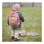 Child with a pink floral backpack facing away on a grassy field, with a small pink lunch box nearby.