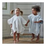 Two joyful toddlers in oversized poncho robes run hand in hand indoors, smiling as they play.