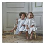 Two young children wearing white robes sit on small wooden chairs, barefoot, laughing together in a bright room with paneled walls.
