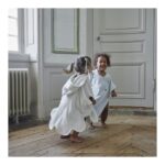 Two young girls in white robes dancing happily indoors near a paneled white door and radiator on a wooden floor