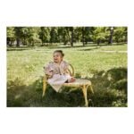 Baby in a beige robe sits barefoot on a wicker chair in a sunlit park.