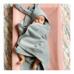 Baby wrapped in a gray towel with a hood featuring bunny ears lies on a pink surface.