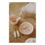 Baby mealtime scene with pink bow‑patterned bowls, cup, spoon, and forks on a rustic wooden table; pink dotted chair padding in the background