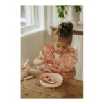 Young girl in a pink floral dress cuts a small cake with a knife at a wooden table.