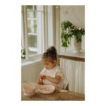 Child with a topknot sits at a wooden table, playing with pink dishware in a sunlit kitchen by a window.