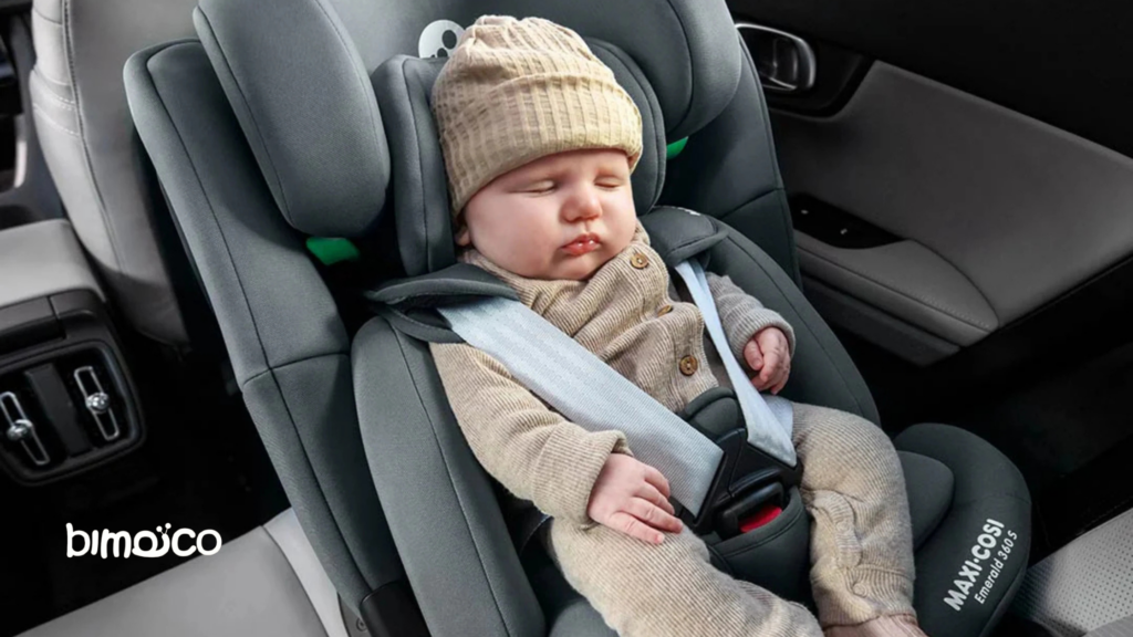 Baby sleeping in a gray infant car seat, wearing a tan knit hat and beige outfit, strapped in with a seatbelt inside a car.