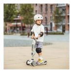 Child wearing a white helmet and protective pads rides a three-wheeled scooter in a sunny urban park.