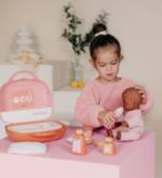 Young girl in a pink sweater feeds a baby doll with a toy bottle at a pink play table with a nurse kit nearby