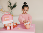 Young girl in a pink sweater changes a baby doll on a pink table with toy bottles and a pink lunchbox nearby