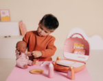 Child playing with a pink baby nurse toy set, feeding a doll at a pink play table.