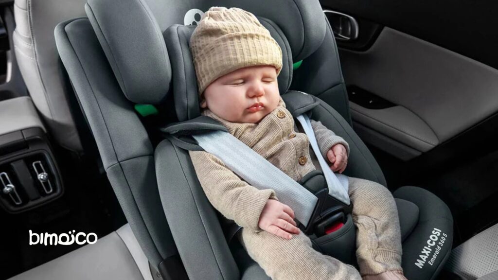 Baby sleeping in a gray infant car seat, wearing a tan knit hat and beige outfit, strapped in with a seatbelt inside a car.