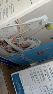 Baby smiling in a colorful infant swing seat mounted on a white frame, shown on product packaging.
