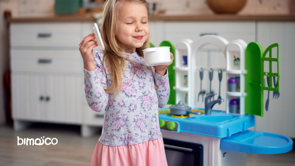 A young girl in a floral shirt and pink skirt holds a white pot and lid in a colorful play kitchen, smiling with eyes closed.