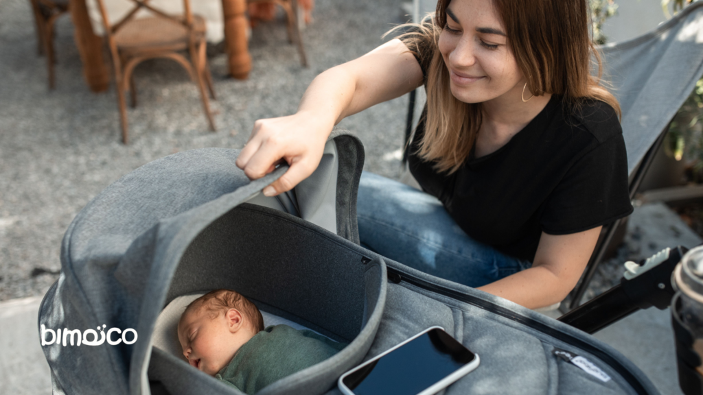 Woman smiling while adjusting a baby in a gray infant car seat/stroller outdoors; a smartphone lies nearby on the seat.