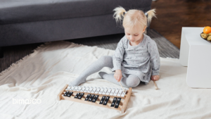 Young girl with blonde pigtails sits on a rug, playing a wooden xylophone indoors with a side table and fruit nearby.