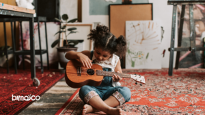 Young girl sits on a patterned red rug, plucking a small wooden guitar in an art studio.