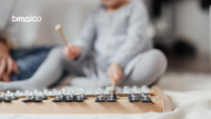 Out-of-focus image of a child playing a wooden peg-and-marble game, with a white 'bimoCo' logo visible on the left.