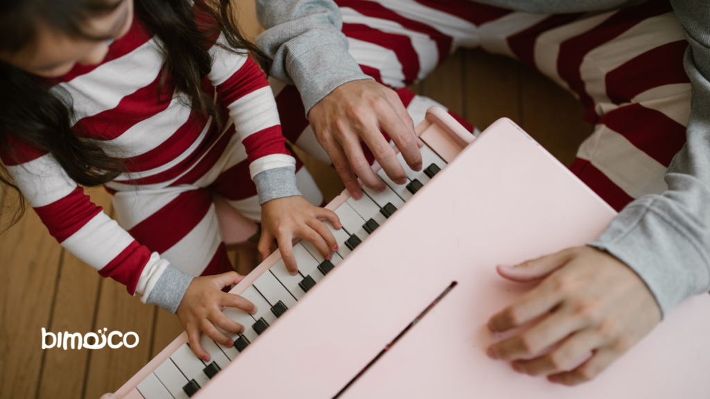 Several hands, including a child's, press the white and black keys of a pink keyboard on a wooden floor, with red-and-white striped clothing in the scene.