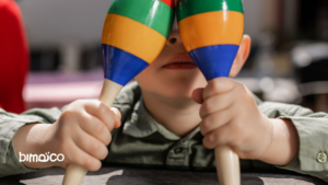 Child playfully covering their eyes with a rainbow-colored mask-like toy, gripping wooden sticks.
