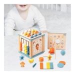 Baby on a white surface playing with a wooden activity cube featuring a clock and colorful bead abacus, surrounded by assorted toys.