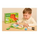 Child coloring in a wipe-clean activity book beside a green dinosaur-themed box labeled 'Wipe-Clean Work Book' with markers on a light table.