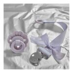 Lavender silicone pacifier with flower-shaped guard and a matching lavender pacifier clip with a fabric ribbon, resting on a crumpled white background (Elodie branding visible).