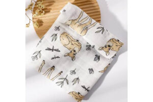 White cloth napkin with a playful print of rabbits, scissors, and leaves, folded on a round wooden board.