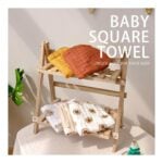 Wooden tiered rack holds folded baby towels in orange, mustard, and sunflower-patterned designs, with a beige backdrop and product title 'Baby Square Towel' visible on the right.