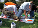 Group of friends bending over a colorful Twister mat on a sunny park lawn.