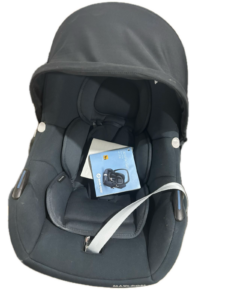 Black infant car seat with extended canopy, padded interior and visible harness straps; a blue instruction booklet tucked inside the seat.