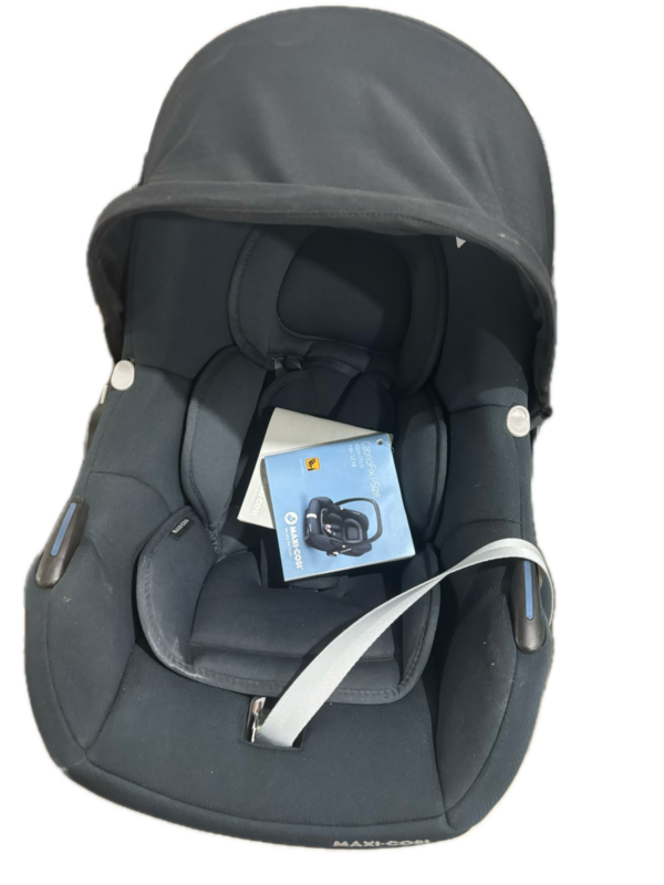 Black infant car seat with extended canopy, padded interior and visible harness straps; a blue instruction booklet tucked inside the seat.