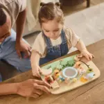 Young girl in denim overalls playing with a wooden sensory puzzle on a table, an adult nearby guiding her.