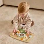 Child sits on a carpet, playing with a wooden busy-board filled with shapes, beads, and colorful components.
