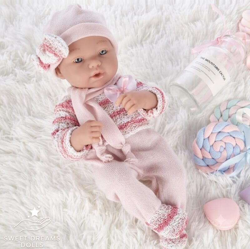 Soft-pink knitted baby doll outfit lying on a white fluffy rug, surrounded by pastel toys and a clear bottle prop (Sweet Dreams Dolls).
