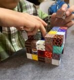 Child in a plaid shirt builds a mosaic of Minecraft texture blocks on a table, placing a block onto a cube structure with colored tiles.