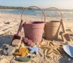 Two pastel buckets with small shovels and assorted sand molds on a sunny beach, ready for play.