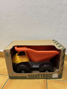 Orange and yellow toy dump truck in an open cardboard box labeled GIANT TRUCK, sitting on a tiled floor.