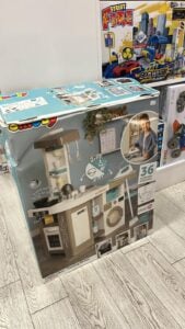 Box of a Smoby pretend kitchen playset with stove, sink, washing machine, and shelves; shows a child using it and notes 36 accessories.