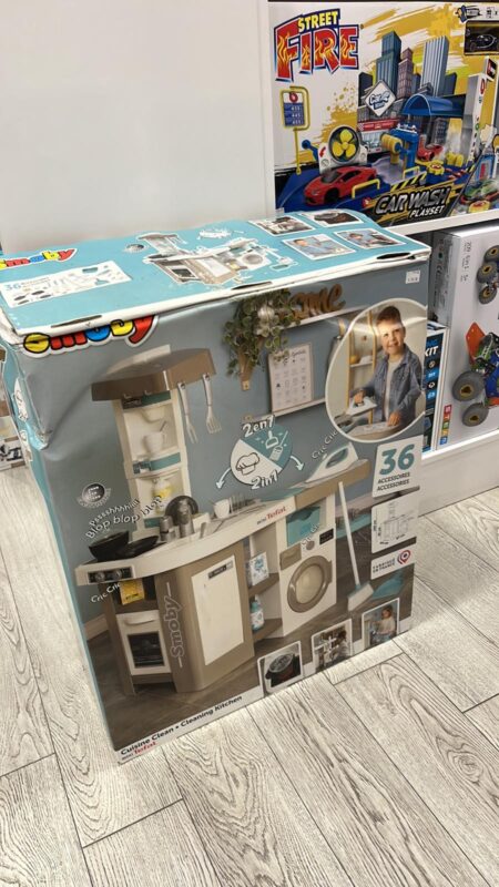 Box of a Smoby pretend kitchen playset with stove, sink, washing machine, and shelves; shows a child using it and notes 36 accessories.