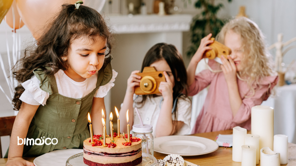 Three children at a birthday party with a cake and lit candles; one girl leans in to blow them out as others hold wooden toy cameras in the background.