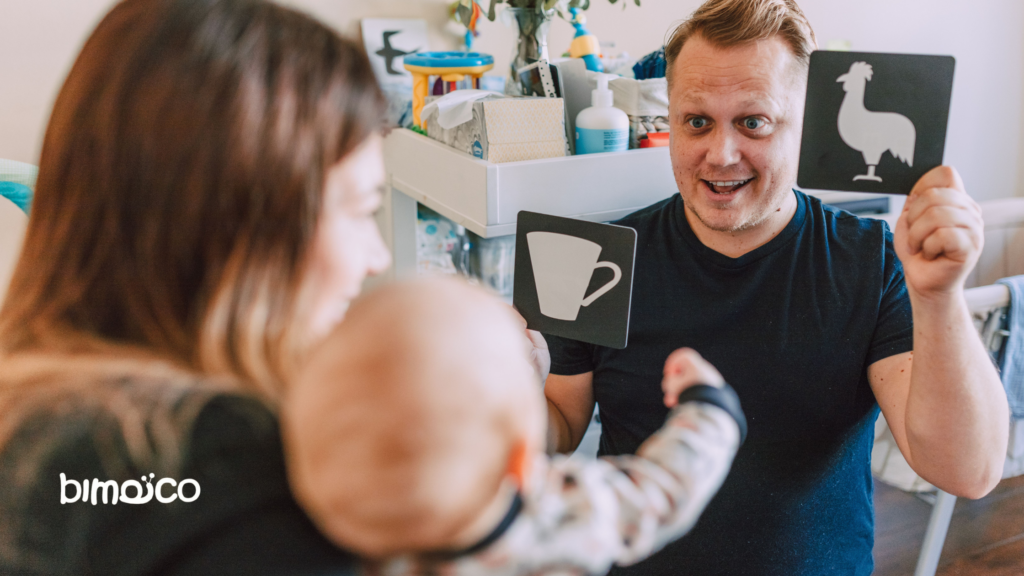 Man in a dark shirt shows two black cards with white icons—a coffee mug and a llama—to a baby held by a woman in the foreground.