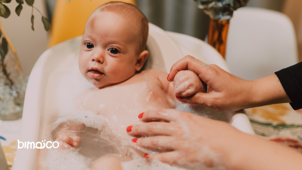 Baby in a white bath tub being washed with soapy water, gentle hands supporting and washing the shoulders.