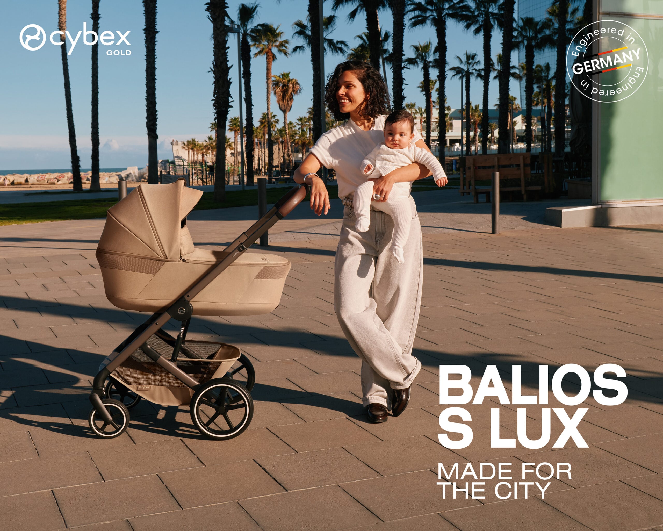 Mother carries a baby in a beige stroller along a palm-lined promenade by the beach; Cybex Gold branding visible on the left.